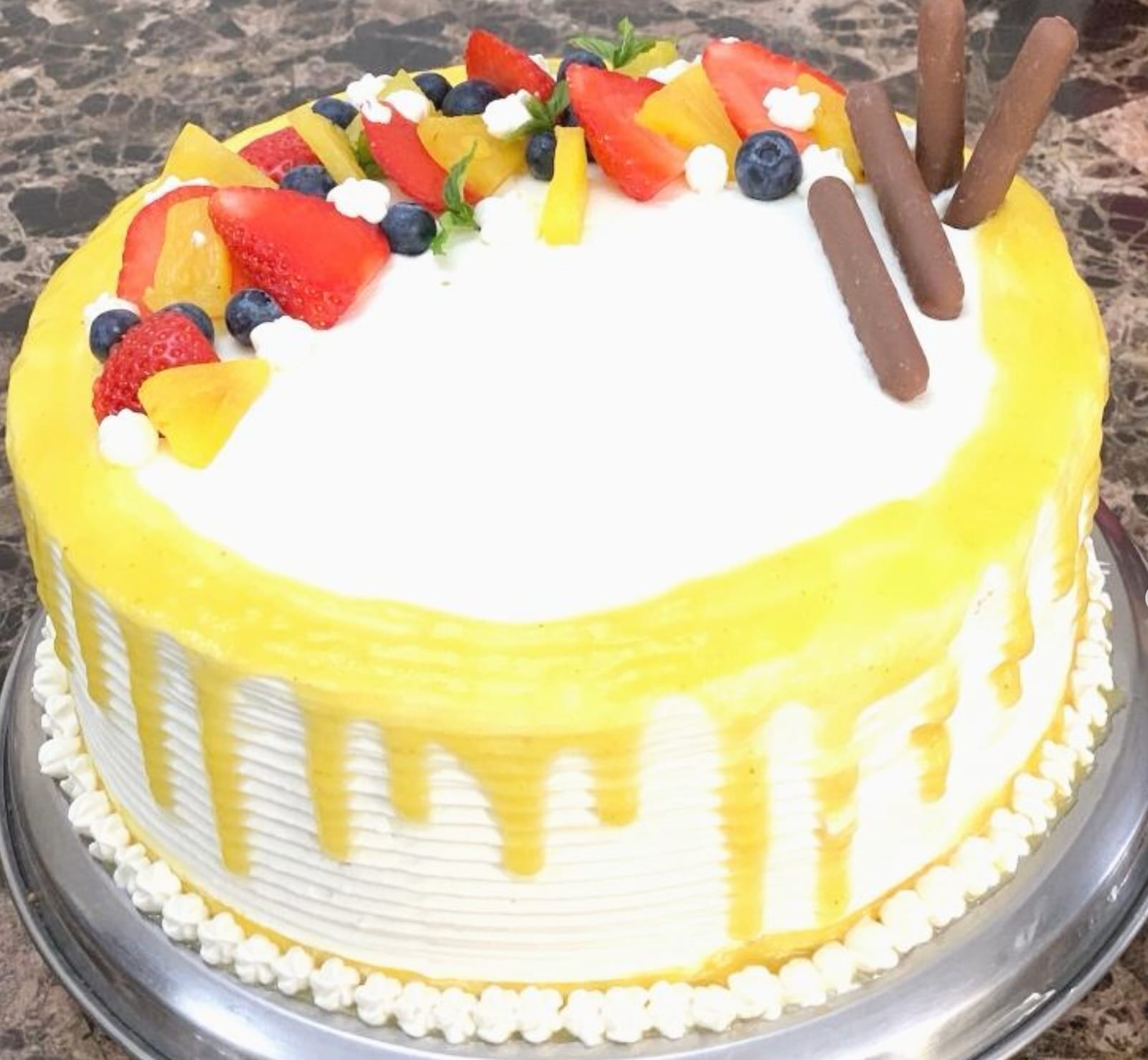Round cream-frosted cake with a pale yellow drip down the sides, topped with strawberries, blueberries, pineapple and kiwi and three chocolate cigar rolls.