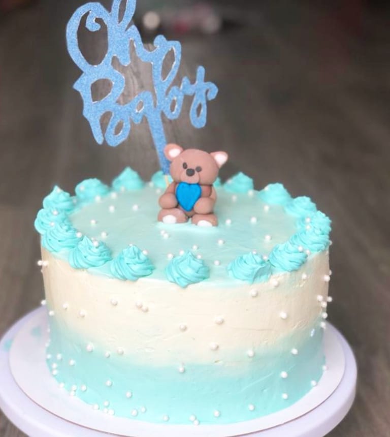 Single-tier cake shading from pale blue at the top down to cream at the base, dotted with sugar pearls, with a piped blue crown, a fondant teddy bear and a glittered Oh Baby topper.