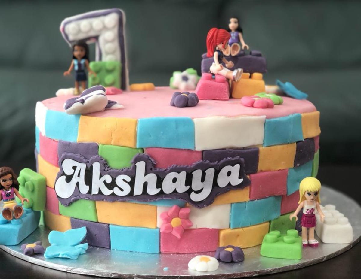 Round cake covered in coloured fondant bricks with a child's name piped across the front, a number seven topper and small figurines arranged around the top edge.