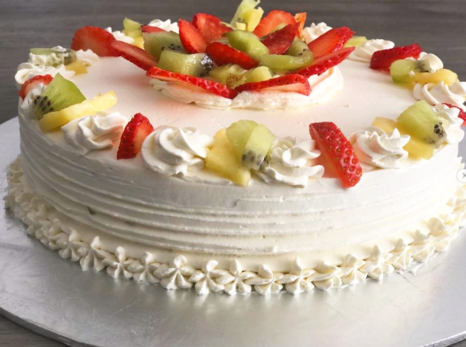 Round cream-frosted cake with a piped shell border around the base and a crown of sliced strawberries, kiwi and pineapple arranged on top.